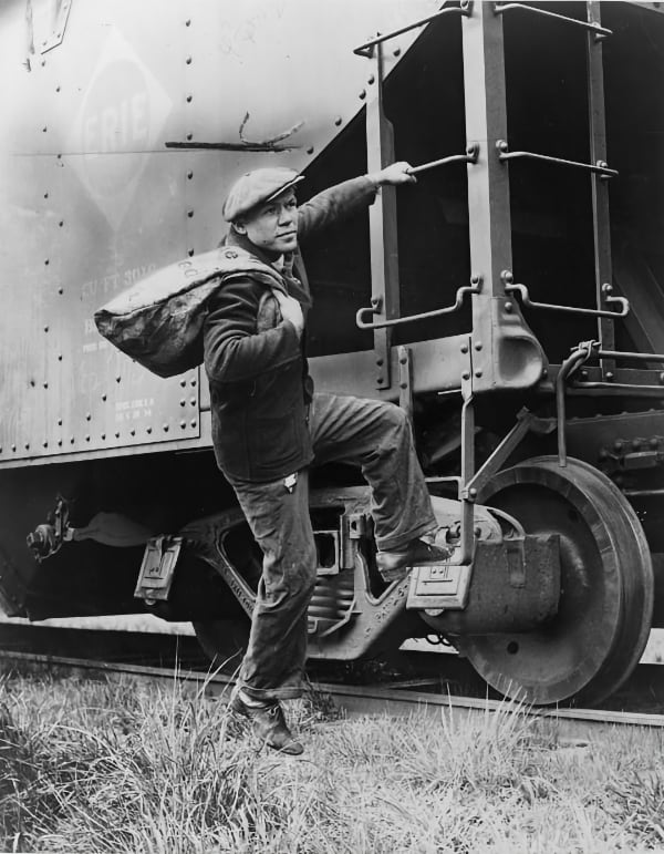 Black and white photo of Lou Ambers with a large bag over his shoulder, mounting the ladder of a train car