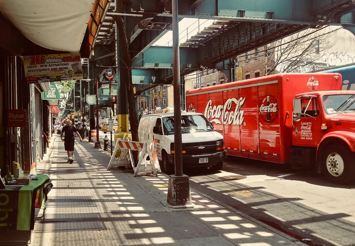 Color photo of a Queens street. The street is under two elevated subway tracks. There is a white van parked on the right side of the street, and a red Coca Cola truck next to the van on the road. A man walks towards the camera on the sidewalk to the right of the road. To his right are many storefronts that appear edge on to the camera, so only certain hanging signs are visible above the sidewalk. One store has a banner with its name, "Zee Wireless Corner," which sells phones. Behind that business is one with a green awning with the words, "Bicycle Sales Repairs," slightly obscured and in white print. Further down the road, it looks like there is construction, as there are construction barrels and barricade visible in the picture. The photo is crowded with so much on this Queens street, and it gives the viewer a claustrophobic feeling.