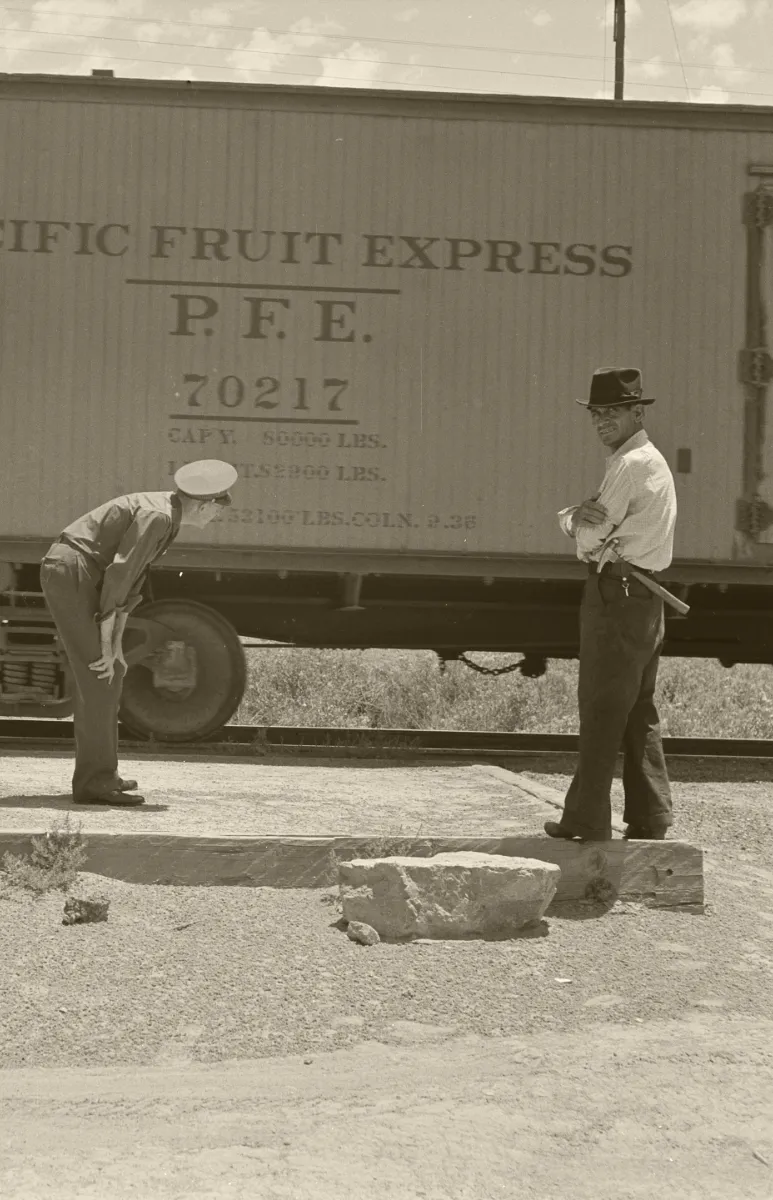 Black and white photo of two railroad workers checking to see if anyone is hiding under a train.